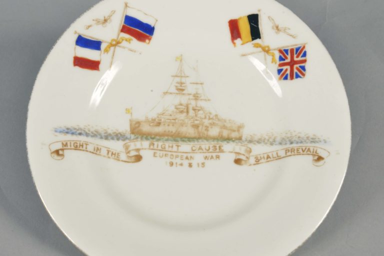 White Grosvenor china World War One Commemorative Plate with a gold rim, made in late 1914. On it are flags of the Allies (France, Russia, Belgium and Britain), a ship and the words: ‘Might in the right cause shall prevail. European War 1914 & 15’. The maker is Jackson & Gosling, England.