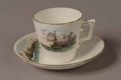 Demi Tasse white porcelain coffee cup and saucer with a gold rim around the top. Front of cup is decorated with a picture of a World War One battleship firing a broadside with painted words: ‘H.M Battleship in action’. Top of saucer is decorated with a picture of a World War One torpedo boat with a white ensign at the jack. Words on the saucer read: ‘H.M Torpedo Boat’.